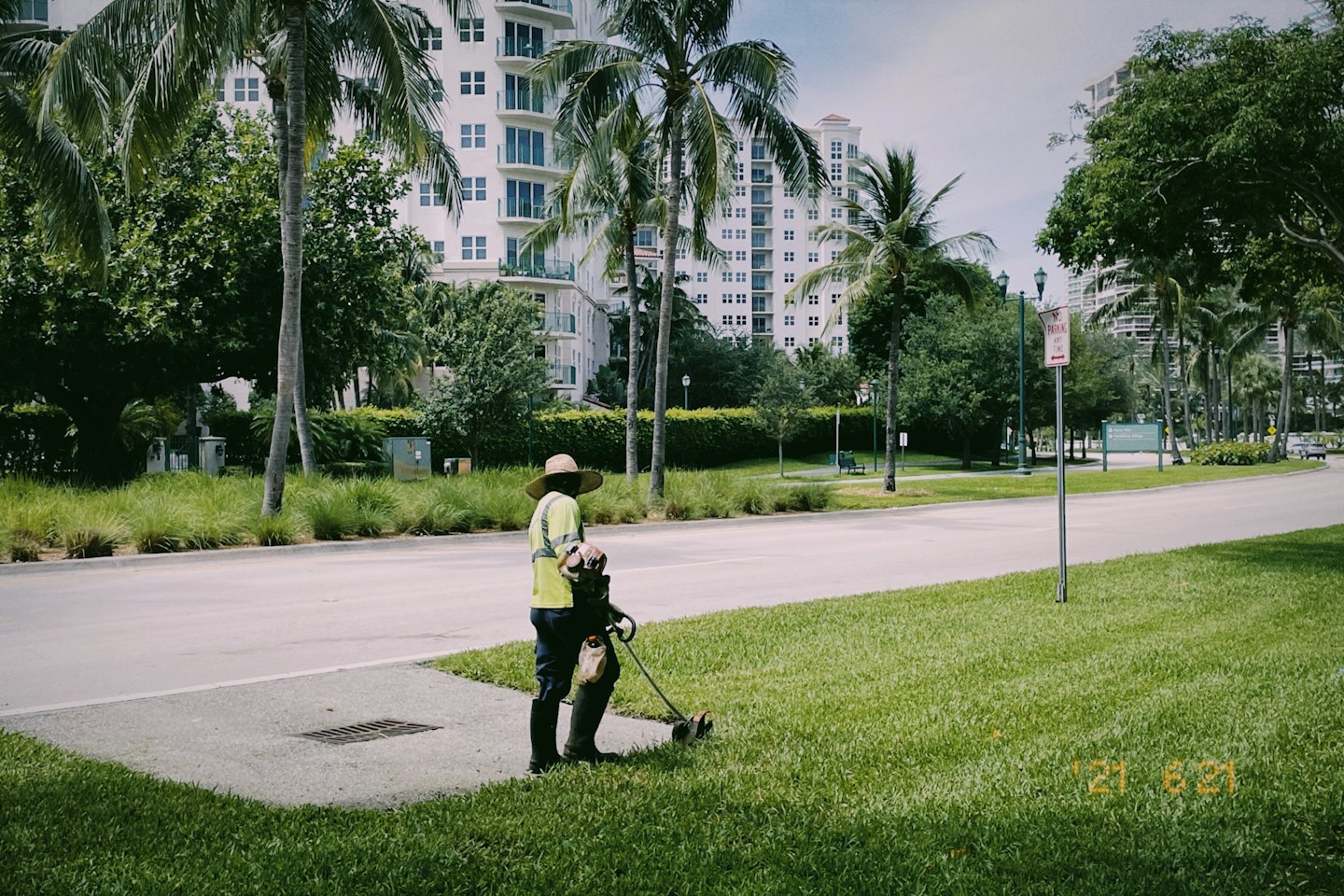 Landscaper trims grass along a sidewalk near tall buildings.